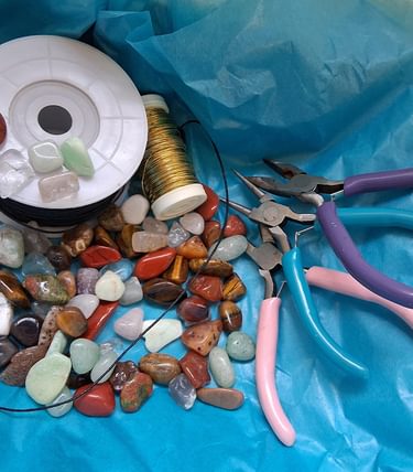 A selection of jewellery-making materials arranged on a turquoise background, including polished sea stones, wire, string, beading tools, and colourful-handled pliers—used in the Seaside Jewellery kids workshop.