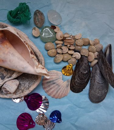 An arrangement of natural and decorative seaside items on a light blue background, including a variety of seashells, pebbles, sea glass, string, and colourful metallic shell-shaped sequins—materials used in the Seaside Shapes kids workshop.