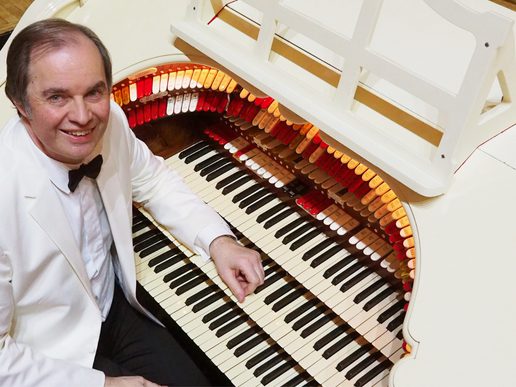 Michael Wooldridge smiling at the console of a white Wurlitzer theatre organ, surrounded by multiple keyboards and rows of colourful stops.