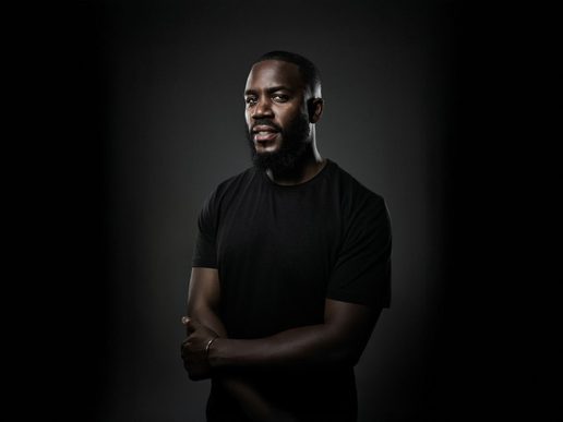 Portrait of comedian Mo Gilligan wearing a black T-shirt, standing with arms loosely crossed against a dark background, looking confidently at the camera.