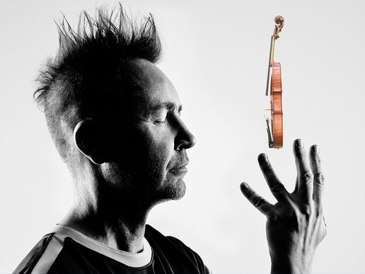 A black-and-white photo of violinist Nigel Kennedy with his eyes closed, holding one hand up as a violin appears to float upright above his fingers against a plain background.