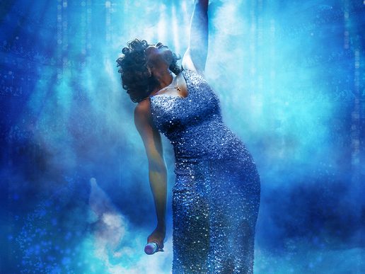 A female-presenting performer in a sparkling blue gown leans backward dramatically while singing into a microphone, one arm raised overhead, bathed in swirling blue light and mist.
