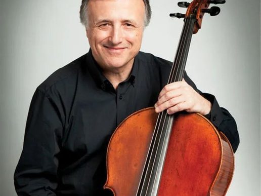 Portrait of cellist Raphael Wallfisch smiling while holding a wooden cello, wearing a black shirt against a plain light grey background.