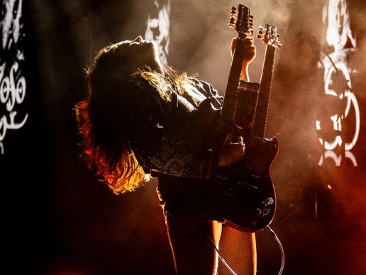 A dramatic moment on stage as a female guitarist leans back while playing a double-neck electric guitar, her hair lit by warm orange stage lights. Smoke and abstract rock-themed visuals surround her, creating an intense rock-concert atmosphere.