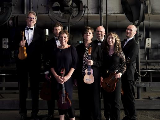 Seven members of The Ukulele Orchestra of Great Britain pose in formal black attire, standing in a semi-industrial setting with large wall-mounted fans and exposed pipes behind them. Each member holds a different type of ukulele, showcasing the variety of instruments used in their performances. The group includes a mix of men and women, with some smiling warmly while others look directly and confidently at the camera. The lighting casts soft shadows, giving the photo a dramatic, performance-ready feel that reflects their unique blend of humour and musicianship.