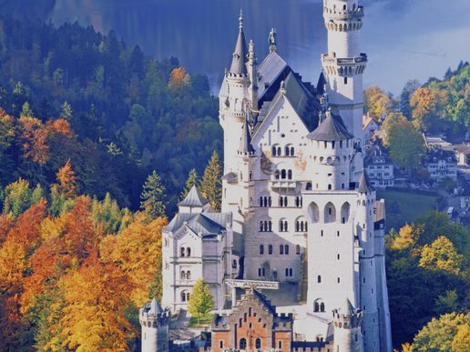 Neuschwanstein Castle rises above a forest of autumn trees in shades of gold, orange and green. Its white stone towers and turrets stand against a backdrop of woodland and a distant lake, creating the appearance of a fairytale castle nestled in the Bavarian landscape.