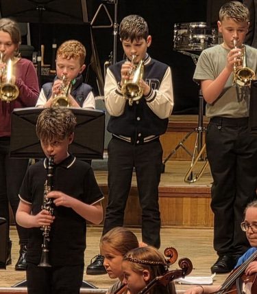 A children's school orchestra performs on stage, with a row of students playing trumpets standing at the back, two students playing clarinets in the middle row, and cellists seated in front, all reading from music stands. A drum kit and timpani are visible in the background.