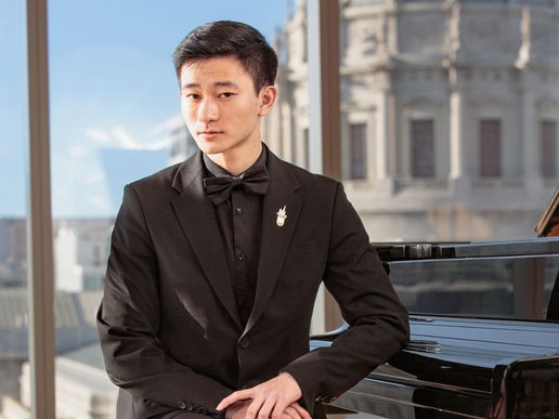 Portrait of pianist Ryan Sheng wearing a black tuxedo and bow tie, standing beside a grand piano in a sunlit room with large windows and a blurred city building in the background.