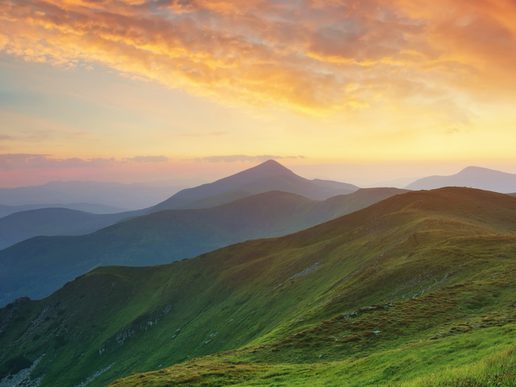 A panoramic view of rolling green mountains stretching into the distance beneath a glowing sunrise. Golden light breaks through dramatic orange clouds, casting a warm glow across the ridges and creating a peaceful, expansive landscape.