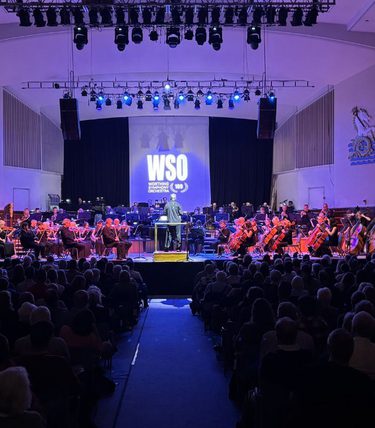 Wide view of the Worthing Symphony Orchestra performing on stage in a large concert hall, with the conductor leading the orchestra under blue stage lighting as a full audience watches.