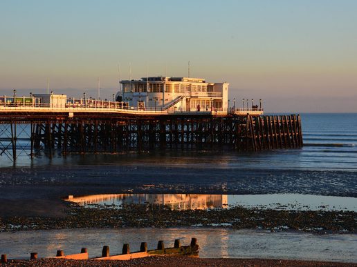 Worthing Pier stretching out over calm sea water at low tide, lit by warm golden sunlight with reflections visible in the wet sand below.