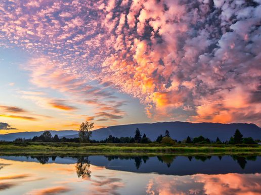 Dramatic sunset sky filled with pink and orange clouds reflected in a calm lake, with dark mountain silhouettes and trees along the horizon.