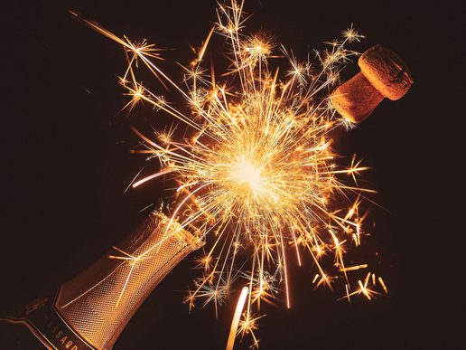 Close-up of a champagne bottle popping open, with the cork flying out and bright golden sparks bursting against a dark background.