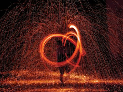 Long-exposure photograph of a performer spinning fire in the dark, creating bright orange arcs and showers of sparks that fill the scene with swirling patterns of light.