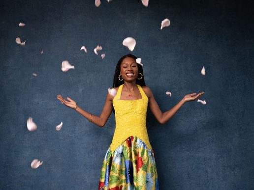Concert pianist Isata Kanneh-Mason smiles with her arms outstretched as pale flower petals fall around her, wearing a yellow top and colourful floral skirt against a dark blue background.
