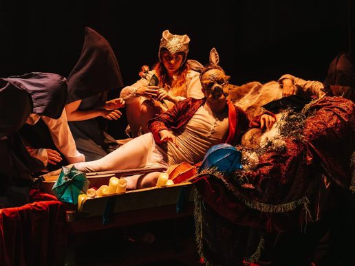 Performers in Flabbergast Theatre’s A Midsummer Night’s Dream gather around a candlelit bed in an atmospheric scene, wearing fantastical costumes and animal-inspired masks.