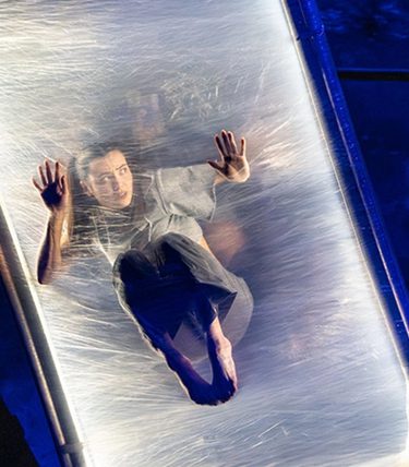 A contemporary circus performer is suspended mid-air inside a transparent vertical frame, appearing to press against a taut, elastic surface. Another performer stabilises the structure on the side, with dramatic blue lighting enhancing the surreal and immersive stage atmosphere.