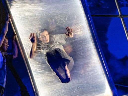 A contemporary circus performer is suspended mid-air inside a transparent vertical frame, appearing to press against a taut, elastic surface. Another performer stabilises the structure on the side, with dramatic blue lighting enhancing the surreal and immersive stage atmosphere.