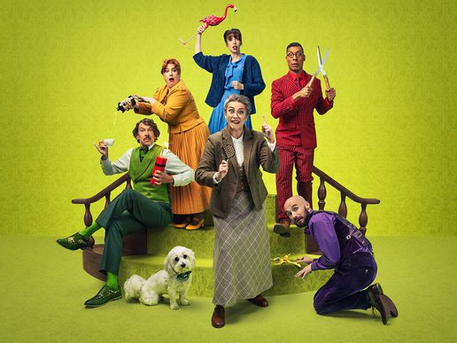A colourful cast of actors pose in a whimsical scene on a bright green set. A woman dressed as a detective stands at the front pointing upward, while characters around her engage in comic antics — one pours tea mid-spill, another holds a flamingo above her head, one brandishes garden shears, and another slips on a banana skin. A small white dog sits calmly at the front. The image promotes the comedy show “Murder, She Didn’t Write.”