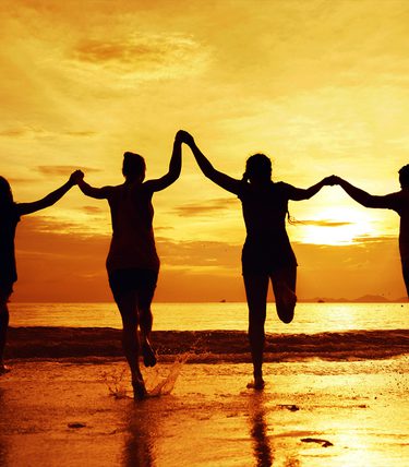Silhouettes of four women holding hands and running joyfully into the sea at sunset. The sky glows with warm shades of orange and gold, reflecting off the water and wet sand. Promotional image for The Worthing Girls, a new concert musical at WTM.