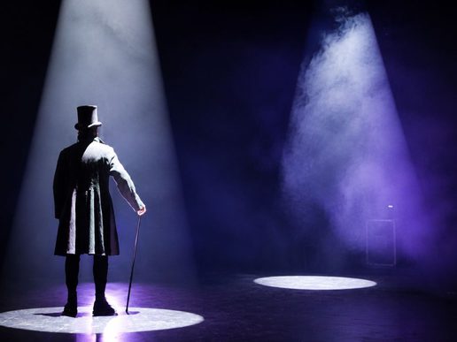 A lone figure in a long Victorian coat and top hat stands on a dark stage holding a cane, illuminated by a bright spotlight and surrounded by theatrical mist, creating a dramatic scene reminiscent of A Christmas Carol.