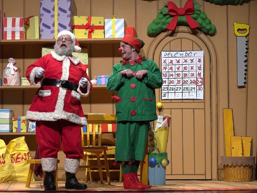 Santa Claus and a cheerful elf stand together on a colourful Christmas workshop stage filled with wrapped presents and festive props. Behind them, a large advent-style calendar on a wooden door shows the days of December being counted down to Christmas. Santa wears a traditional red suit with white trim, while the elf is dressed in bright green with red buttons and curled red shoes. The warm, playful set creates a fun and festive atmosphere for a family Christmas show.