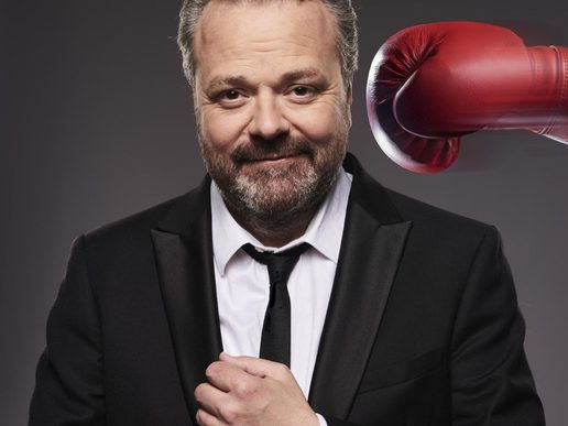 A man in a black suit and tie adjusts his cuff, smiling at the camera, while a red boxing glove is positioned close to the side of his head against a plain dark background.