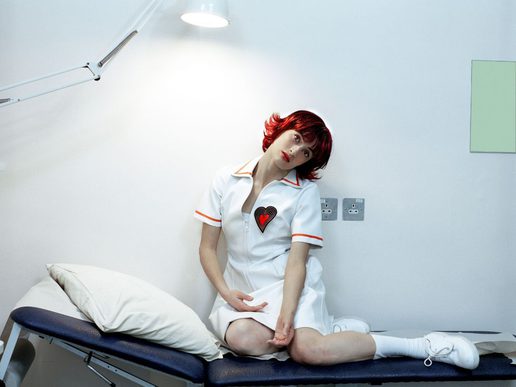 A young woman with short, bright red hair sits on a medical examination couch in a stark white clinical room. She wears a white nurse-style dress with orange trim and a heart-shaped emblem on the chest, along with white socks and trainers. Her head tilts to one side as she gazes thoughtfully towards the camera beneath a large examination lamp, creating a mood that is both clinical and dreamlike. A pillow rests beside her on the couch, while the minimalist surroundings emphasise her solitary pose.