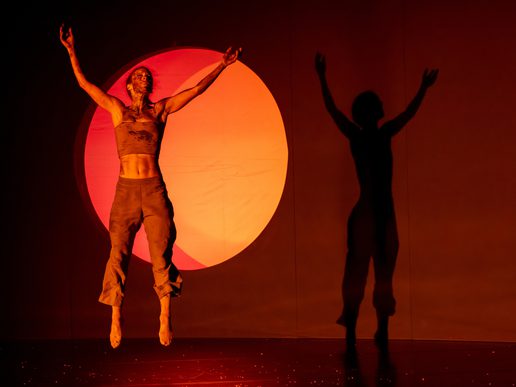 A dancer from Solstice leaps with arms raised in front of a glowing orange circle, casting a dramatic shadow across the red-lit stage.
