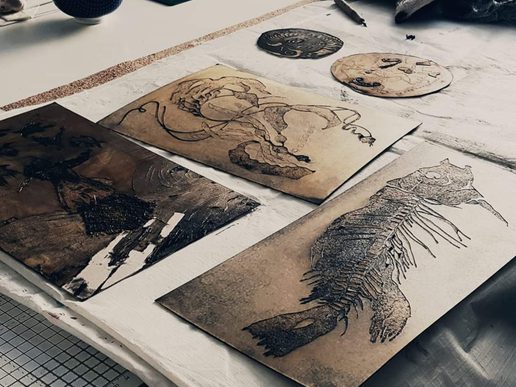 Collagraph printmaking plates and prints featuring detailed animal designs, including a fish and other textured illustrations, arranged on a workshop table.