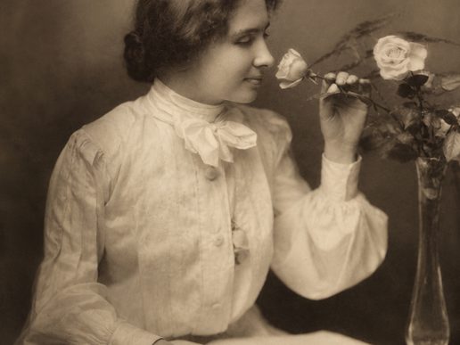 A sepia-toned portrait of Helen Keller as a young woman, seated in profile while gently smelling a rose. She wears a high-necked white blouse with a large bow at the collar, and a vase of blooming roses sits beside her, creating a calm and reflective scene.