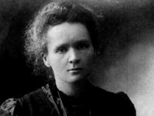 Black-and-white portrait of physicist and chemist Marie Curie looking directly at the camera. She wears a dark high-necked dress, with her hair swept back in a loose updo. The softly lit studio portrait highlights her calm, thoughtful expression against a plain, dark background.