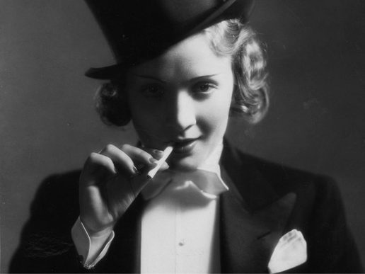 Black-and-white portrait of Marlene Dietrich wearing a top hat and tailored tuxedo jacket with a white shirt and bow tie. She faces the camera with a confident, slightly playful expression, holding a cigarette near her lips. Dramatic lighting creates soft shadows across her face, highlighting her curled hair and refined features against a dark background.
