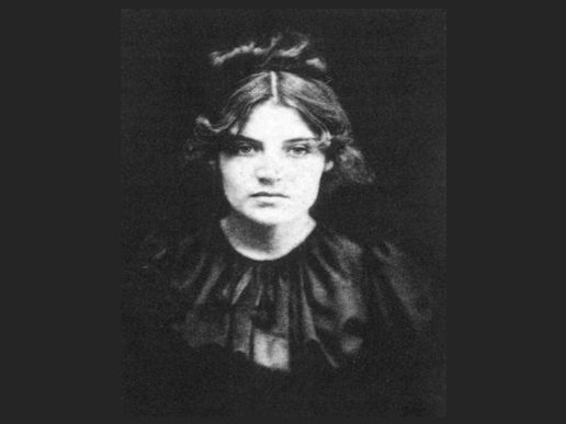 A black-and-white portrait of Suzanne Valadon as a young woman. She faces the camera with a calm, serious expression, wearing a dark dress with a gathered neckline. Her shoulder-length hair is parted in the middle and softly curled, set against a plain dark background.