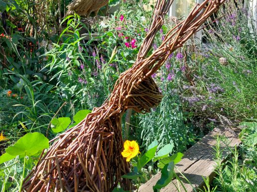 Handwoven willow hare sculpture surrounded by colourful flowers and lush green plants in a sunlit garden.