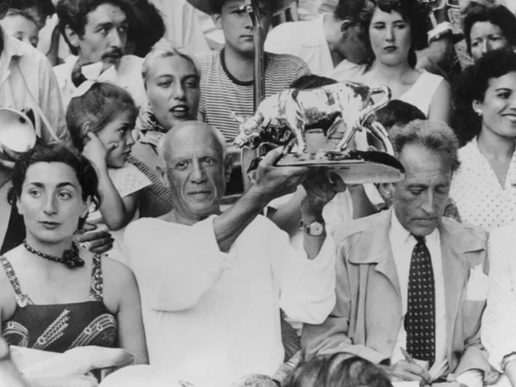 Black-and-white photograph of artist Pablo Picasso holding a metallic bull trophy above his head while seated in a crowded audience. To his right, writer Jean Cocteau sits looking down and writing, and to his left a woman in a patterned dress looks away from the camera. The scene is lively, with many people gathered closely together.