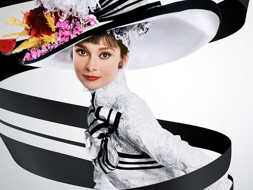Portrait of Audrey Hepburn wearing an elegant white lace dress and a wide-brimmed black-and-white striped hat adorned with pink flowers, looking directly at the camera. A flowing black ribbon wraps around her against a clean white background, creating a striking, stylised composition.