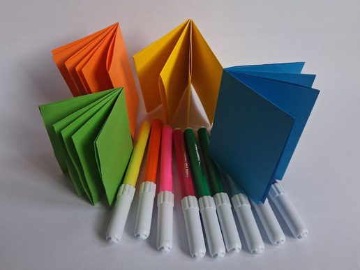 Four small folded paper booklets in bright colours (green, orange, yellow, and blue) arranged in a row, with a set of colourful felt-tip pens laid out in front on a white background.