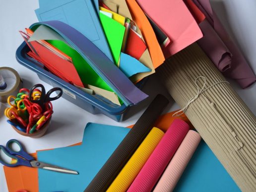 Assorted colourful craft supplies on a table, including stacks of bright paper in a plastic tray, rolls of corrugated card, scissors, tape, and a pot of multicoloured paper clips.