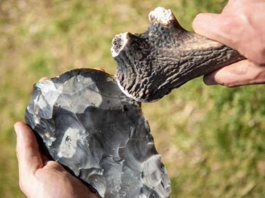 Hands holding a piece of flint and an antler tool, demonstrating traditional flint knapping outdoors.