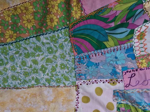 Close-up of a handmade patchwork quilt made from colourful fabric pieces stitched together with visible decorative embroidery. Floral prints in green, blue, yellow and pink sit alongside bold abstract patterns and polka dots, highlighting the quilt's handcrafted detail and vibrant mix of vintage-style fabrics.