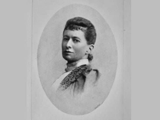 Black-and-white oval portrait of a young Victorian woman looking directly at the camera. She has dark hair swept up in a neat updo and wears a high-collared dress with a patterned shoulder detail. The formal studio portrait is set against a plain light background.
