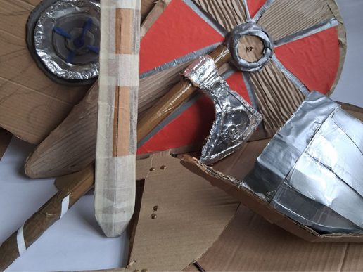 Handmade cardboard props arranged together, including a sword, axe, shields, and helmet, crafted from corrugated card, tape, and foil with simple painted details.