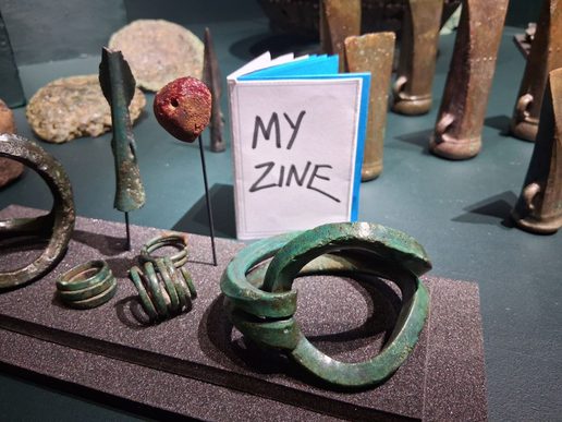 Small handmade booklet titled “My Zine” displayed among museum artefacts, including metal rings, bracelets, and other aged objects arranged on a dark display surface.