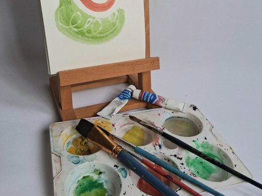 Watercolour painting setup with a small canvas on a wooden easel, tubes of paint, brushes, and a paint palette with mixed colours on a white surface.