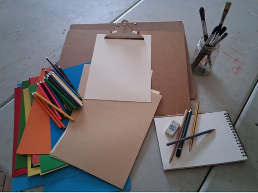 Art materials arranged on a table, including coloured pencils, coloured paper, drawing boards, a sketchbook, eraser and a jar of paintbrushes.