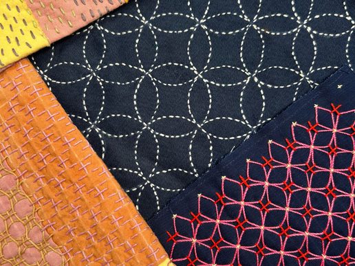 Close-up of decorative stitched fabrics, showing geometric embroidery patterns in white, red, and yellow thread on dark blue and warm-toned cloth pieces.