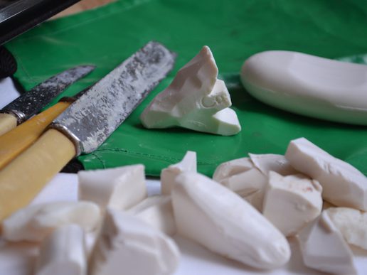 Close-up of white soap pieces being carved on a green cutting mat, with carving knives and a partially shaped piece of soap.