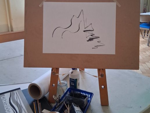 A simple black line drawing of a jug and angular still-life forms displayed on an easel, with drawing and printmaking materials arranged on the table below.