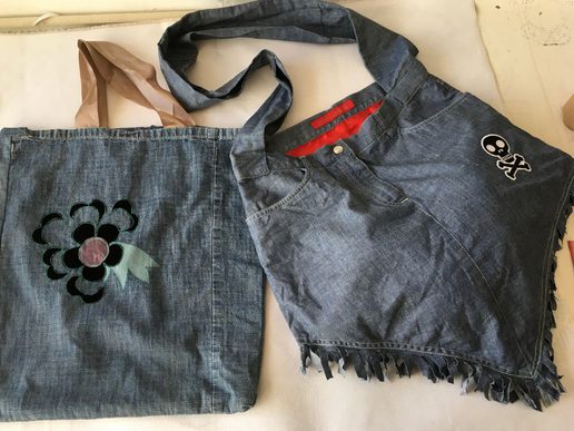 Upcycled denim items laid flat, including a tote bag with a flower appliqué and a cropped denim top with a skull patch and fringed hem.